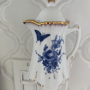 May include: A white porcelain plug-in night light shaped like a miniature pitcher with a lid. The surface features a blue floral pattern and a blue butterfly. The rim of the pitcher and the handle are accented with gold trim, and the lid has a gold finial.