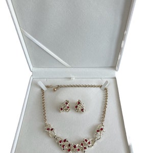 Peut inclure: Un ensemble collier et boucles d'oreilles rouge et or dans un coffret à bijoux en velours blanc. Le collier présente une chaîne délicate avec un motif de pierres rouges serties dans des feuilles d'or. Les boucles d'oreilles sont assorties.