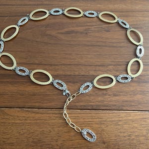 Puede incluir: Un cintur&oacute;n de metal dorado y plateado con eslabones ovalados. El cintur&oacute;n tiene una serie de eslabones ovalados dorados que se alternan con eslabones ovalados plateados. El cintur&oacute;n est&aacute; sobre una superficie de madera.