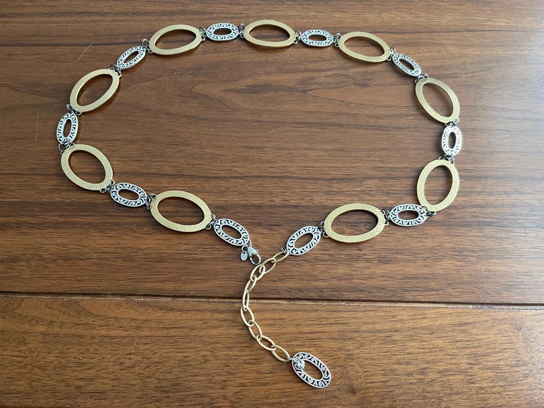 Puede incluir: Un cintur&oacute;n de metal dorado y plateado con eslabones ovalados. El cintur&oacute;n tiene una serie de eslabones ovalados dorados que se alternan con eslabones ovalados plateados. El cintur&oacute;n est&aacute; sobre una superficie de madera.