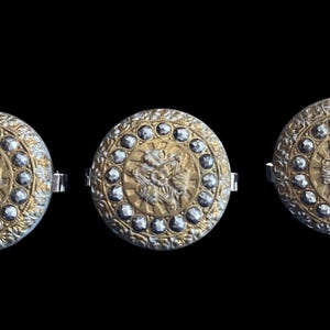 May include: Three decorative, circular buttons with a gold central design of a flower and a silver-coloured outer ring. Each button is adorned with small, clear, faceted stones. The buttons have a silver-coloured clasp on the back.