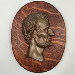 May include: An oval-shaped wall plaque featuring a bronze-toned metal relief profile of Abraham Lincoln. The metallic bust is mounted onto a dark brown wooden base with a visible, textured wood grain pattern throughout the oval surface.