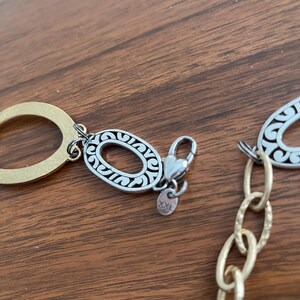 Puede incluir: Primer plano de un collar dorado y plateado. El collar presenta eslabones ovalados con intrincados dise&ntilde;os de filigrana plateada. Un cierre en forma de coraz&oacute;n conecta la cadena. El fondo es una superficie de madera.