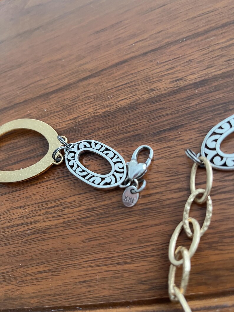 Puede incluir: Primer plano de un collar dorado y plateado. El collar presenta eslabones ovalados con intrincados dise&ntilde;os de filigrana plateada. Un cierre en forma de coraz&oacute;n conecta la cadena. El fondo es una superficie de madera.