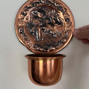 May include: A circular copper wall planter featuring a relief design of a person sitting in a wooded area with a dog. The piece has a polished copper finish with dark patina accents in the recessed areas and a smooth, rounded copper basin.