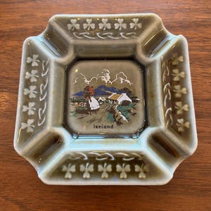 May include: A square ceramic ashtray with a glossy olive green glaze. The rim features embossed white shamrock patterns. The centre depicts a colourful illustration of a person in a field near a cottage with the text Ireland printed below.