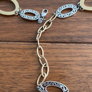 Puede incluir: Un cintur&oacute;n dorado y plateado con eslabones ovalados. El cintur&oacute;n presenta un dise&ntilde;o decorativo dentro de los eslabones plateados y una extensi&oacute;n de cadena. El cierre es plateado. El cintur&oacute;n se muestra sobre una superficie de madera.