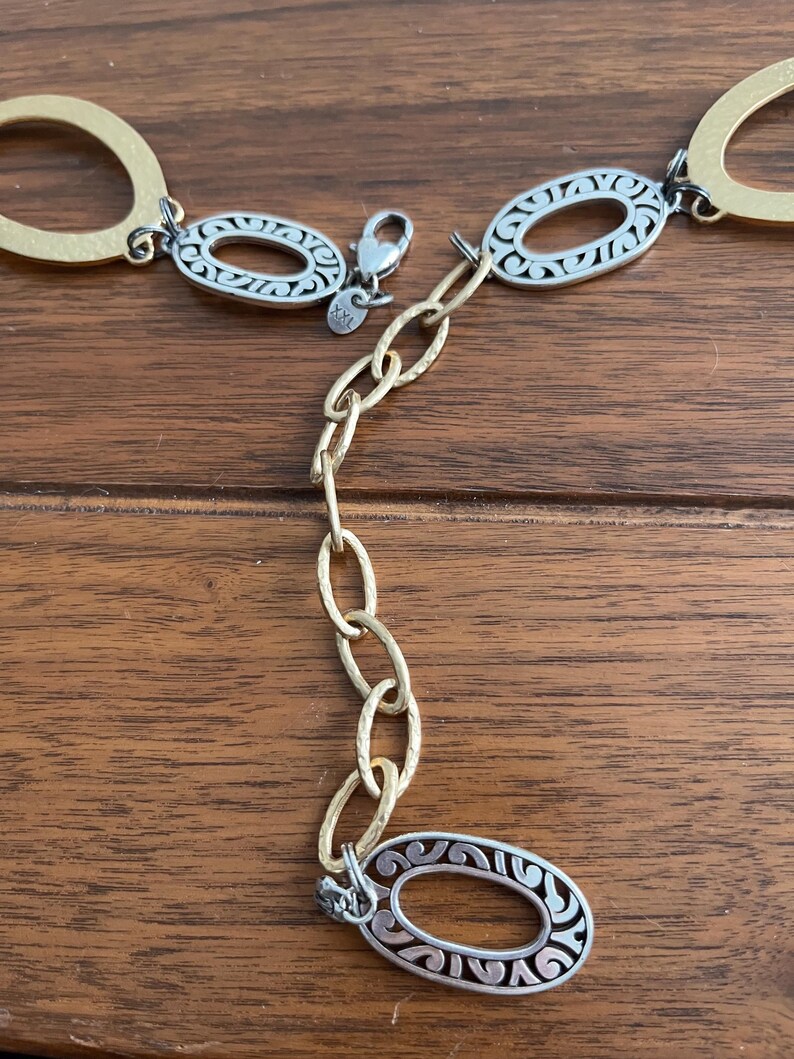 Puede incluir: Un cintur&oacute;n dorado y plateado con eslabones ovalados. El cintur&oacute;n presenta un dise&ntilde;o decorativo dentro de los eslabones plateados y una extensi&oacute;n de cadena. El cierre es plateado. El cintur&oacute;n se muestra sobre una superficie de madera.