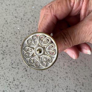 May include: A circular cabinet knob featuring a white floral pattern within a gold-coloured frame. The knob has a central gold dot and is held in a hand.