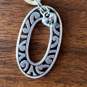 Puede incluir: Colgante ovalado plateado con un dise&ntilde;o ornamental y arremolinado, conectado a una cadena dorada. El colgante tiene un peque&ntilde;o detalle en forma de coraz&oacute;n en la parte superior. La joya descansa sobre una superficie de madera.