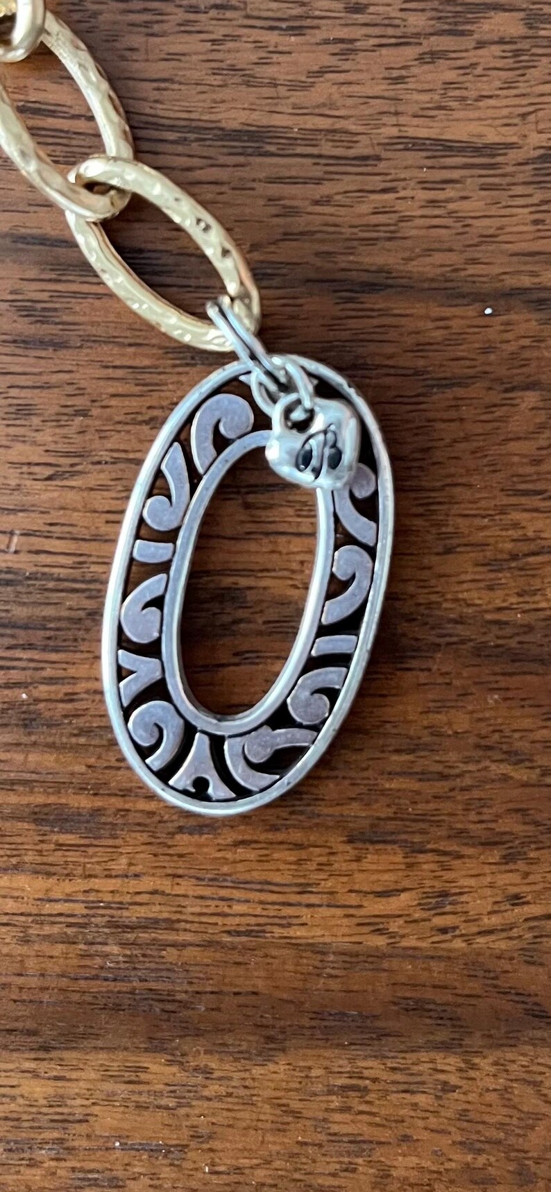 Puede incluir: Colgante ovalado plateado con un dise&ntilde;o ornamental y arremolinado, conectado a una cadena dorada. El colgante tiene un peque&ntilde;o detalle en forma de coraz&oacute;n en la parte superior. La joya descansa sobre una superficie de madera.