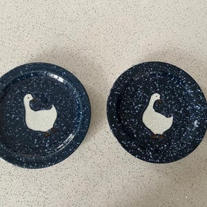 May include: Two navy blue plates with a speckled pattern and a white goose design. The plates are round and likely made of ceramic or porcelain. Suitable for serving food or as decorative items.