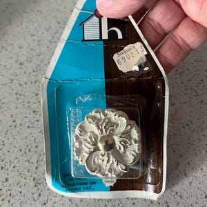 Peut inclure: Un bouton de meuble blanc en forme de fleur ornée, dans son emballage d'origine. L'emballage est en forme de maison avec des sections bleues et marron. L'étiquette indique "Simpson's 6902T3" et "CANADA".