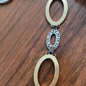Puede incluir: Un collar dorado y plateado con eslabones ovalados. El collar presenta eslabones ovalados martillados de color dorado y eslabones ovalados de filigrana plateada, conectados por peque&ntilde;os eslabones negros. La joya se exhibe sobre una superficie de madera.