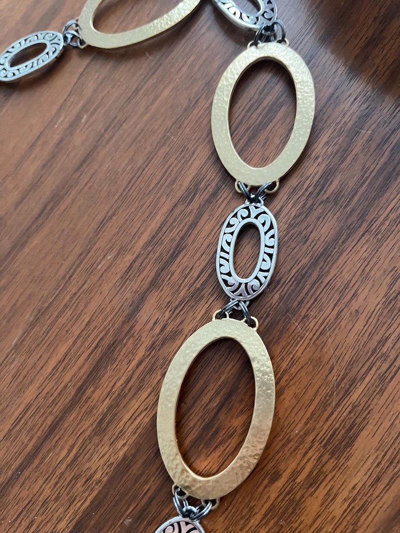 Puede incluir: Un collar dorado y plateado con eslabones ovalados. El collar presenta eslabones ovalados martillados de color dorado y eslabones ovalados de filigrana plateada, conectados por peque&ntilde;os eslabones negros. La joya se exhibe sobre una superficie de madera.