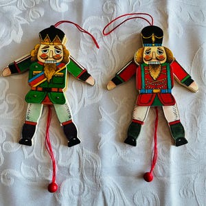 Peut inclure: Deux jouets en bois en forme de casse-noisette avec des détails peints en rouge, vert et blanc. Les casse-noisettes sont habillés en soldats et ont une ficelle attachée à leurs jambes. La ficelle est attachée à une boule rouge.