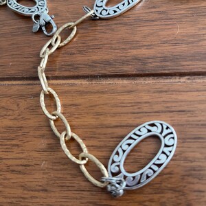 Puede incluir: Un cintur&oacute;n de metal plateado y dorado con eslabones ovalados. Los eslabones ovalados tienen un dise&ntilde;o decorativo de filigrana. El cintur&oacute;n tiene un cierre de mosquet&oacute;n y una cadena de eslabones ovalados dorados.