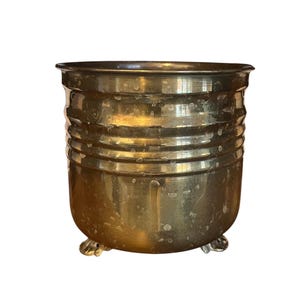 May include: A vintage brass planter, cylindrical with horizontal ribbed detailing. It stands on three small feet. The brass has a warm, aged patina, giving it an antique appearance. A decorative piece for the home.