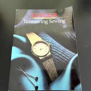 May include: A softcover book titled Singer Sewing Reference Library Timesaving Sewing. The cover features a gold-toned wristwatch with a mesh band resting on blue fabric, accompanied by two straight pins with black heads against a dark background.