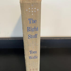 May include: A hardcover book titled "The Right Stuff" by Tom Wolfe. The book's spine is beige with blue lettering and star designs. The publisher's initials, F.S.G., are at the bottom.