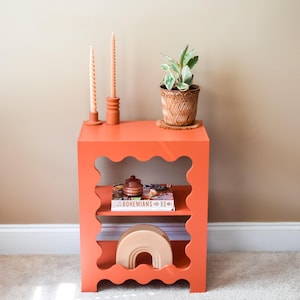 May include: A bright orange side table with scalloped edges and two shelves. The table is decorated with a small plant in a brown pot, two candles, a book titled "The Bohemians", and a decorative object.
