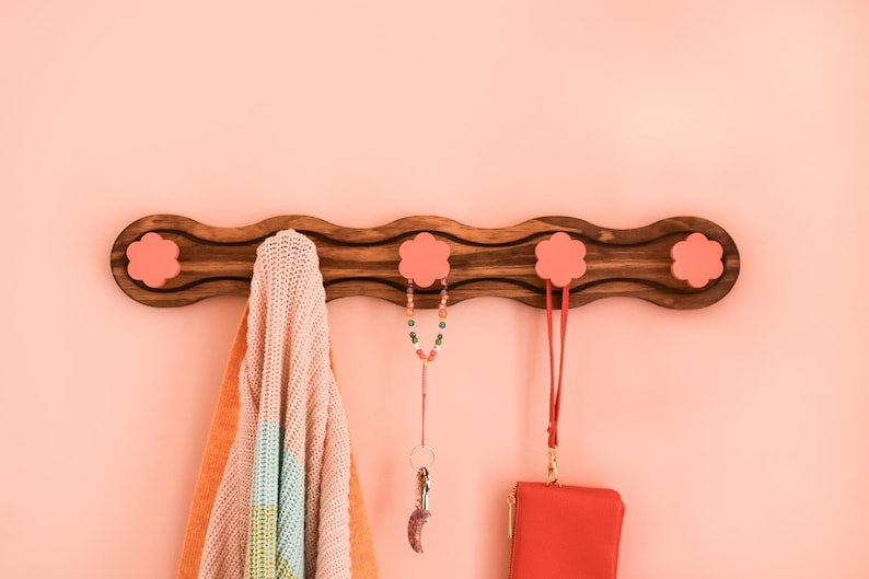 May include: A wooden wall-mounted coat rack with four pink flower-shaped hooks. A multi-colored beaded necklace, a knitted scarf, and a red purse are hanging from the hooks.
