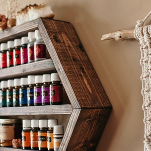 May include: A dark brown, hexagonal wooden shelf filled with essential oil bottles of various colors. The shelf is mounted on a beige wall, with a macrame wall hanging and decorative items nearby. The shelf is designed to display essential oils.