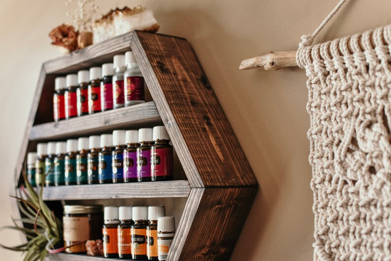 May include: A dark brown, hexagonal wooden shelf filled with essential oil bottles of various colors. The shelf is mounted on a beige wall, with a macrame wall hanging and decorative items nearby. The shelf is designed to display essential oils.