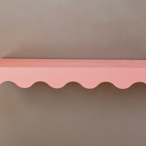 Peut inclure: Une &eacute;tag&egrave;re rose &agrave; bord festonn&eacute; avec une bordure ondul&eacute;e. L'&eacute;tag&egrave;re est fix&eacute;e &agrave; un mur gris clair.
