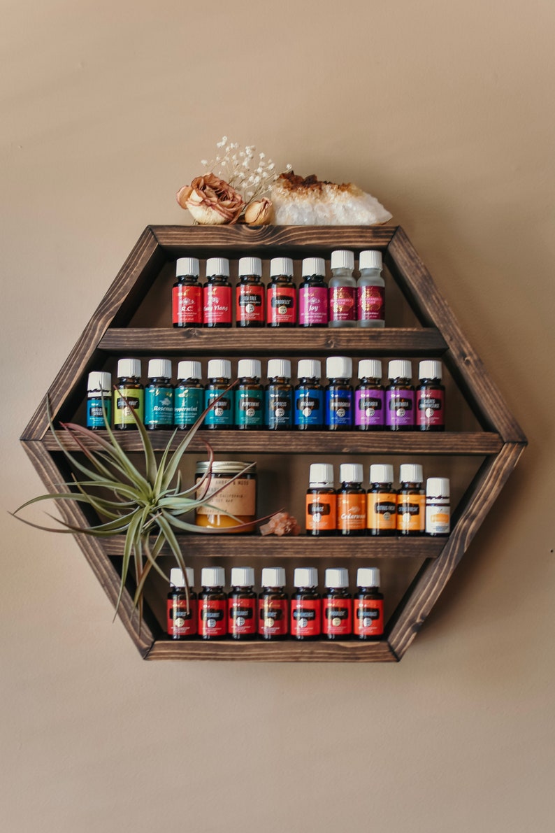 May include: A dark stained, hexagonal wooden shelf displays rows of essential oil bottles with white caps and colorful labels. The shelf is decorated with a dried flower, crystal, and air plant. The oils are organized by color.