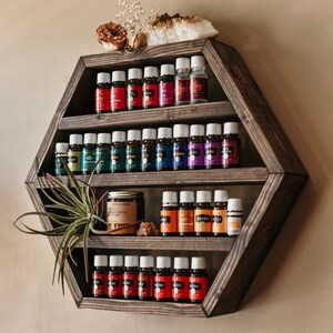 May include: A dark stained, hexagonal wooden shelf displays rows of essential oil bottles with colorful labels. The shelf is decorated with a dried flower, a crystal, and a small air plant. The shelf is mounted on a neutral-colored wall.