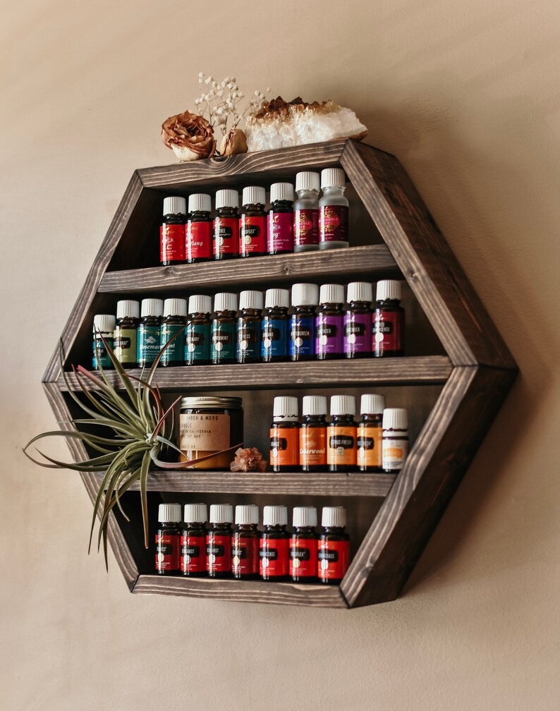 May include: A dark stained, hexagonal wooden shelf displays rows of essential oil bottles with colorful labels. The shelf is decorated with a dried flower, a crystal, and a small air plant. The shelf is mounted on a neutral-colored wall.