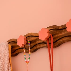 May include: A wooden wall-mounted coat rack with a wavy design and four pink flower-shaped hooks. A colorful beaded necklace and a long pink strap hang from the hooks.