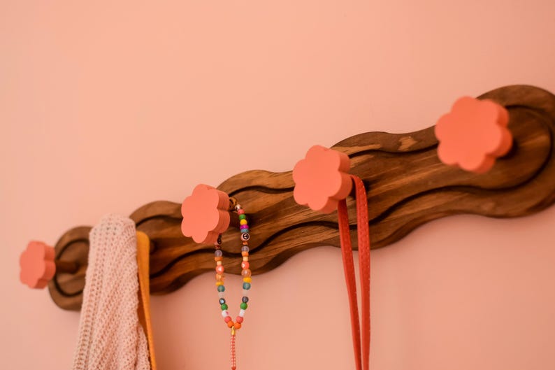 May include: A wooden wall-mounted coat rack with a wavy design and four pink flower-shaped hooks. A colorful beaded necklace and a long pink strap hang from the hooks.
