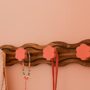 May include: A wooden wall-mounted coat rack with a wavy design. It has four pink flower-shaped hooks. A pink knitted scarf and a colorful beaded necklace are hanging on the hooks.