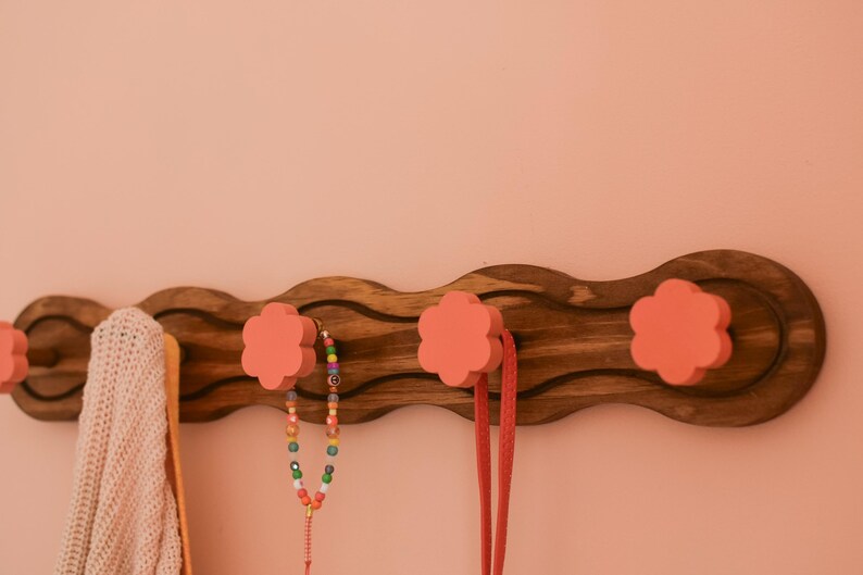 May include: A wooden wall-mounted coat rack with a wavy design. It has four pink flower-shaped hooks. A pink knitted scarf and a colorful beaded necklace are hanging on the hooks.