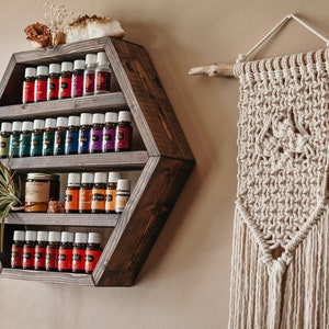 May include: A dark stained, hexagonal wooden shelf displays rows of essential oil bottles with colorful labels. A small air plant and a jar are also on the shelf. To the right, a macrame wall hanging with a natural wood branch.