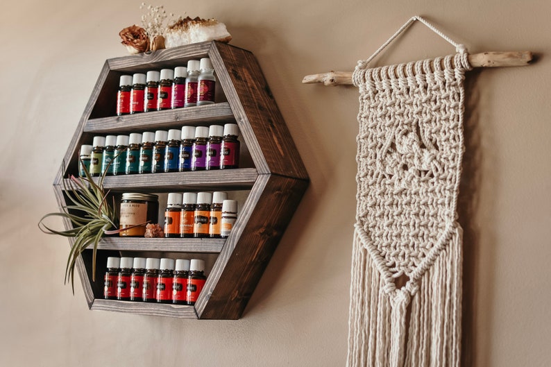 May include: A dark stained, hexagonal wooden shelf displays rows of essential oil bottles with colorful labels. A small air plant and a jar are also on the shelf. To the right, a macrame wall hanging with a natural wood branch.