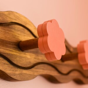 May include: A wooden wall-mounted coat rack with a wavy design. The rack has four hooks with pink flower-shaped knobs.