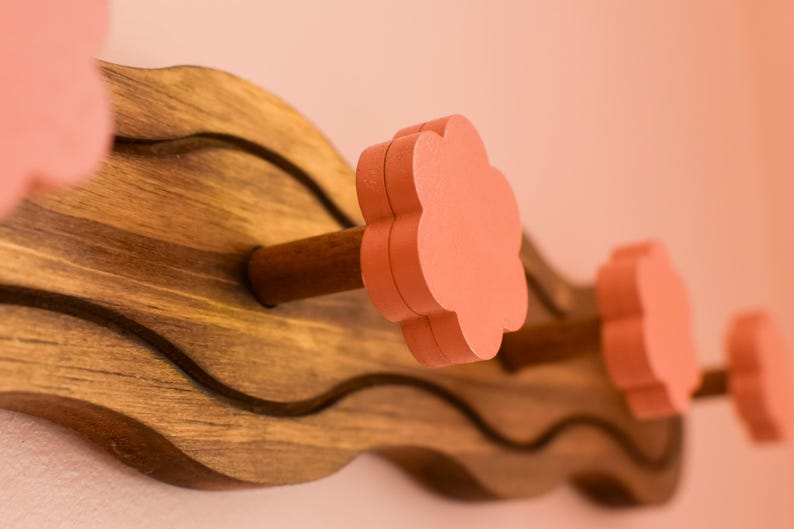 May include: A wooden wall-mounted coat rack with a wavy design. The rack has four hooks with pink flower-shaped knobs.