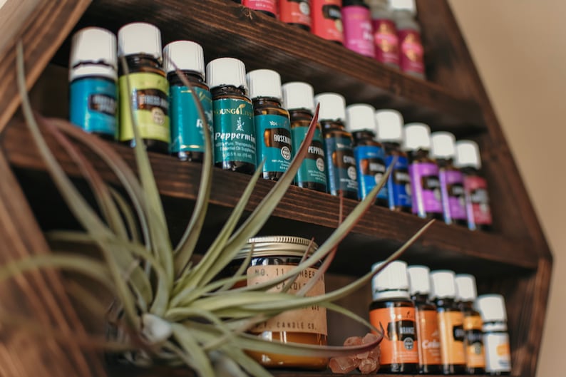 May include: A dark wooden shelf displays various essential oil bottles with white caps and colorful labels. A jar candle and air plant are in the foreground. The shelf is filled with aromatherapy products.