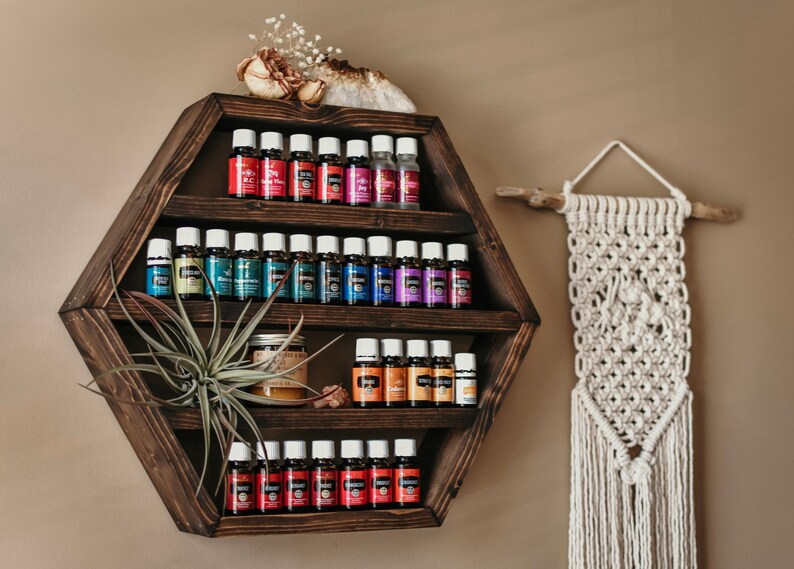 May include: A dark brown, hexagonal wooden shelf filled with small essential oil bottles in various colors. A decorative macrame wall hanging and a small air plant are also visible. The shelf is mounted on a neutral-colored wall.