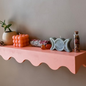 Peut inclure: Une &eacute;tag&egrave;re rose avec un bord festonn&eacute;, d&eacute;cor&eacute;e de divers objets, dont une plante en pot, des bougies, un fagot de sauge et une petite bouteille en verre. L'&eacute;tag&egrave;re est fix&eacute;e &agrave; un mur gris.