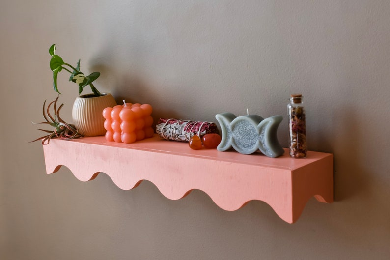 Peut inclure: Une &eacute;tag&egrave;re rose avec un bord festonn&eacute;, d&eacute;cor&eacute;e de divers objets, dont une plante en pot, des bougies, un fagot de sauge et une petite bouteille en verre. L'&eacute;tag&egrave;re est fix&eacute;e &agrave; un mur gris.