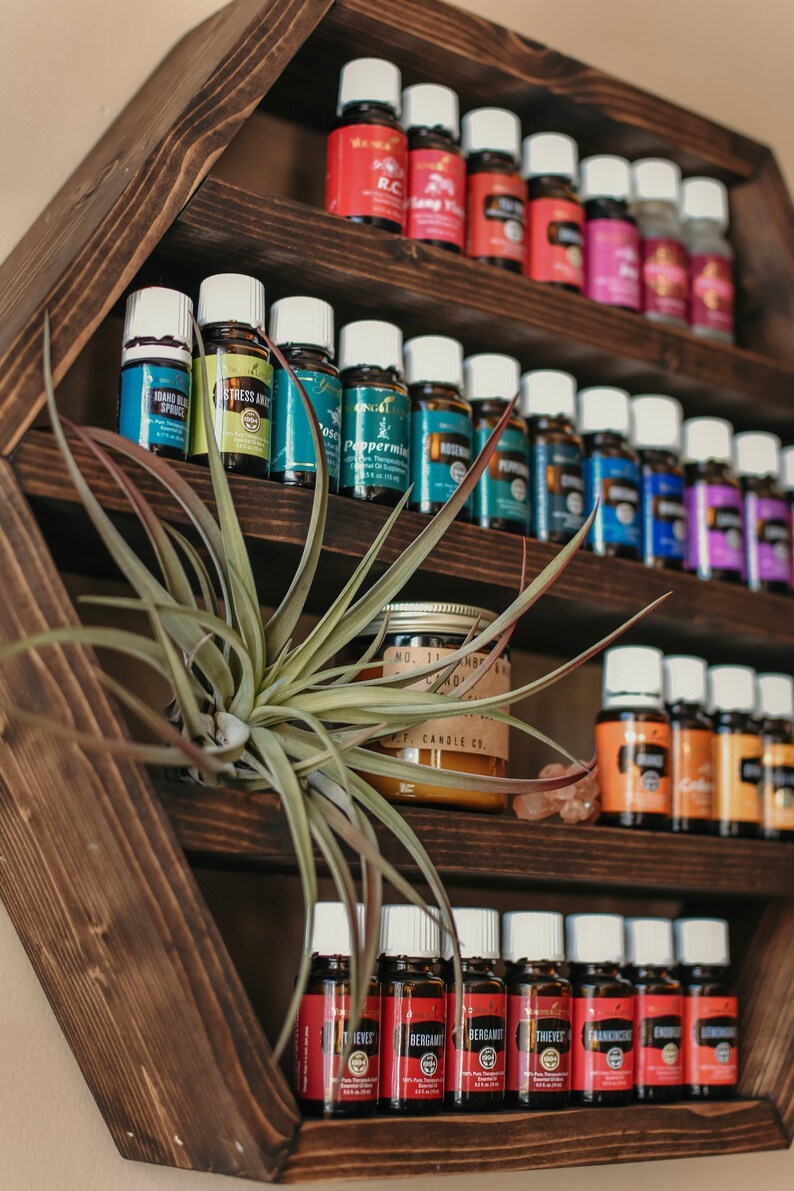 May include: A dark brown, hexagonal wooden shelf displaying numerous essential oil bottles with white caps and colorful labels. A small air plant and a candle are also on the shelf. The oils are organized by type.
