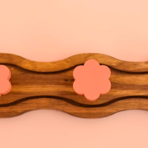 May include: A wooden wall-mounted coat rack with a wavy design. It has three pink flower-shaped hooks.