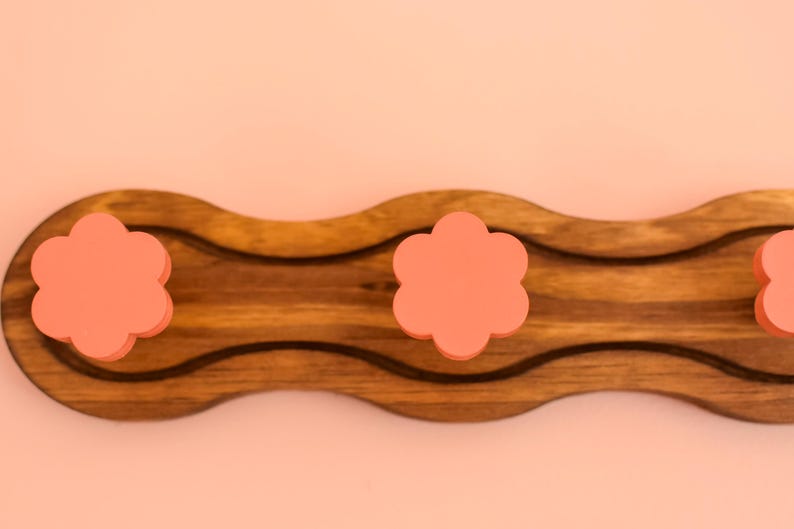 May include: A wooden wall-mounted coat rack with a wavy design. It has three pink flower-shaped hooks.