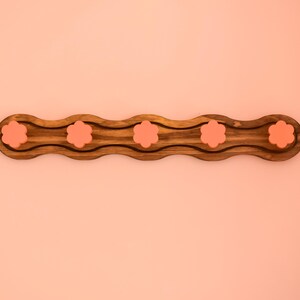 May include: A wooden tray with a wavy design and five pink flower-shaped indentations. The tray is perfect for holding small items like jewelry or keys.