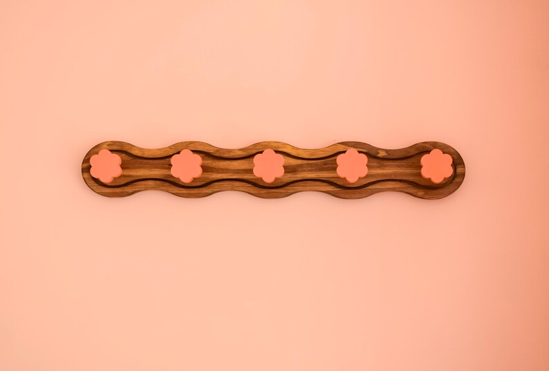 May include: A wooden tray with a wavy design and five pink flower-shaped indentations. The tray is perfect for holding small items like jewelry or keys.