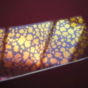 May include: A rectangular piece of material with a pattern of yellow shapes on a purple background. The material has a reflective, metallic appearance, with a gradient of colors from yellow to purple.